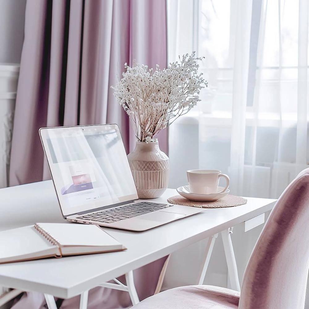 Lilac + Mint customer support desk.  A cozy home office scene with a white desk, lavender curtains, a laptop, a vase of dried flowers, a cup and saucer, and an open notebook on a pink chair.