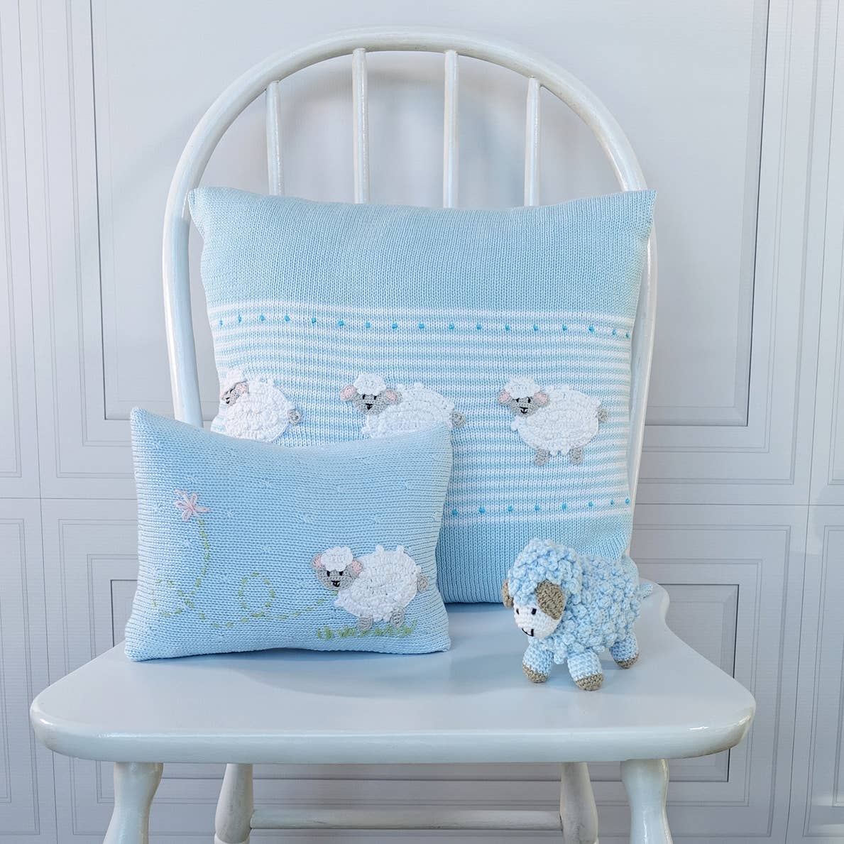 Two light blue crocheted throw pillows with white sheep designs, displayed on a white wooden chair next to a small blue and white sheep plush toy.