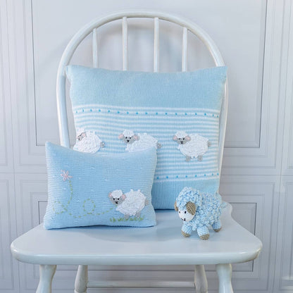 Two light blue crocheted throw pillows with white sheep designs, displayed on a white wooden chair next to a small blue and white sheep plush toy.